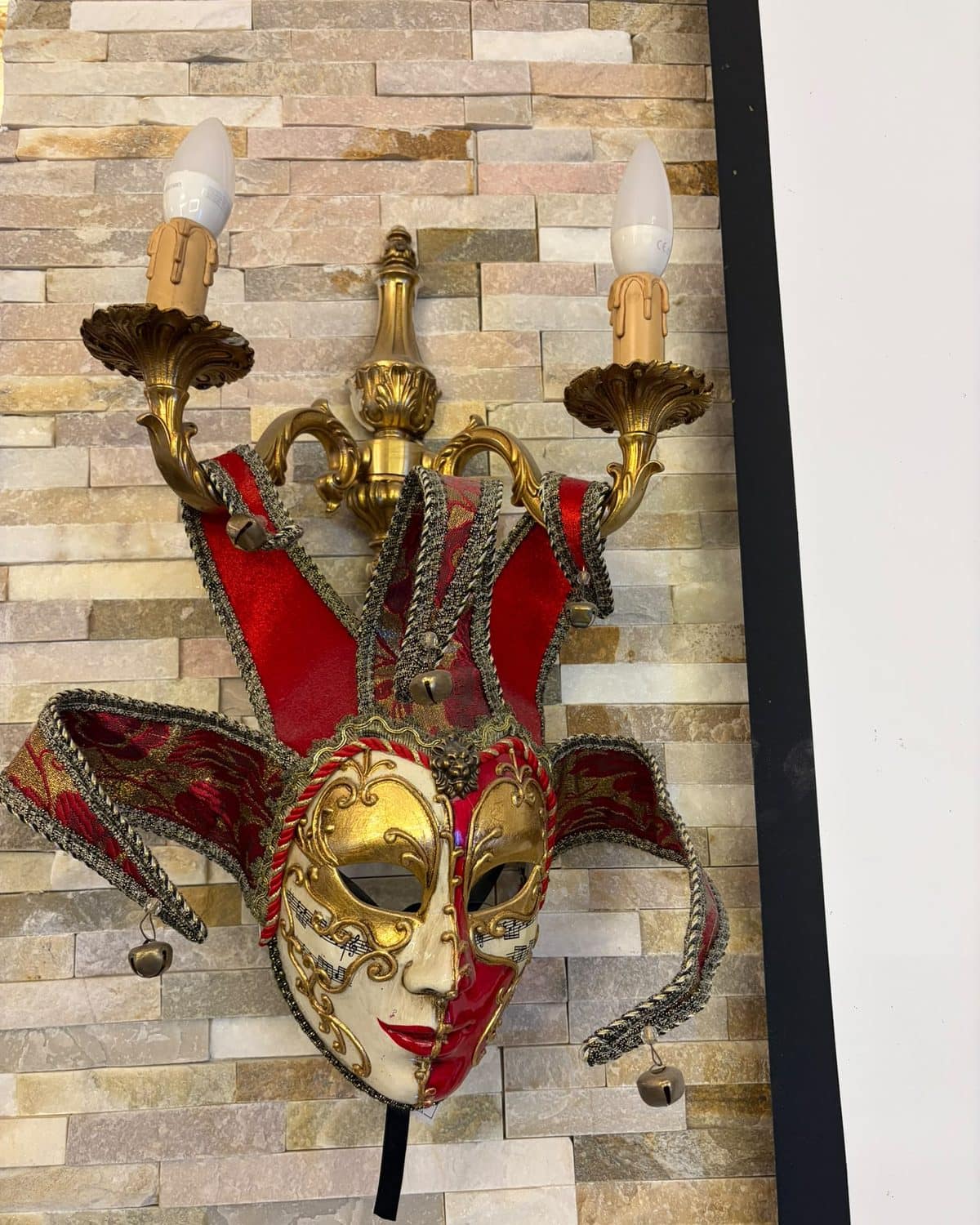 Masque vénitien de bouffon, rouge et or, accroché sous une applique en laiton.