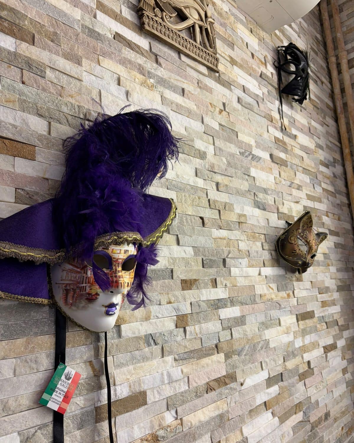Masque vénitien à plumes violettes, accroché au mur de pierre claire de la salle.