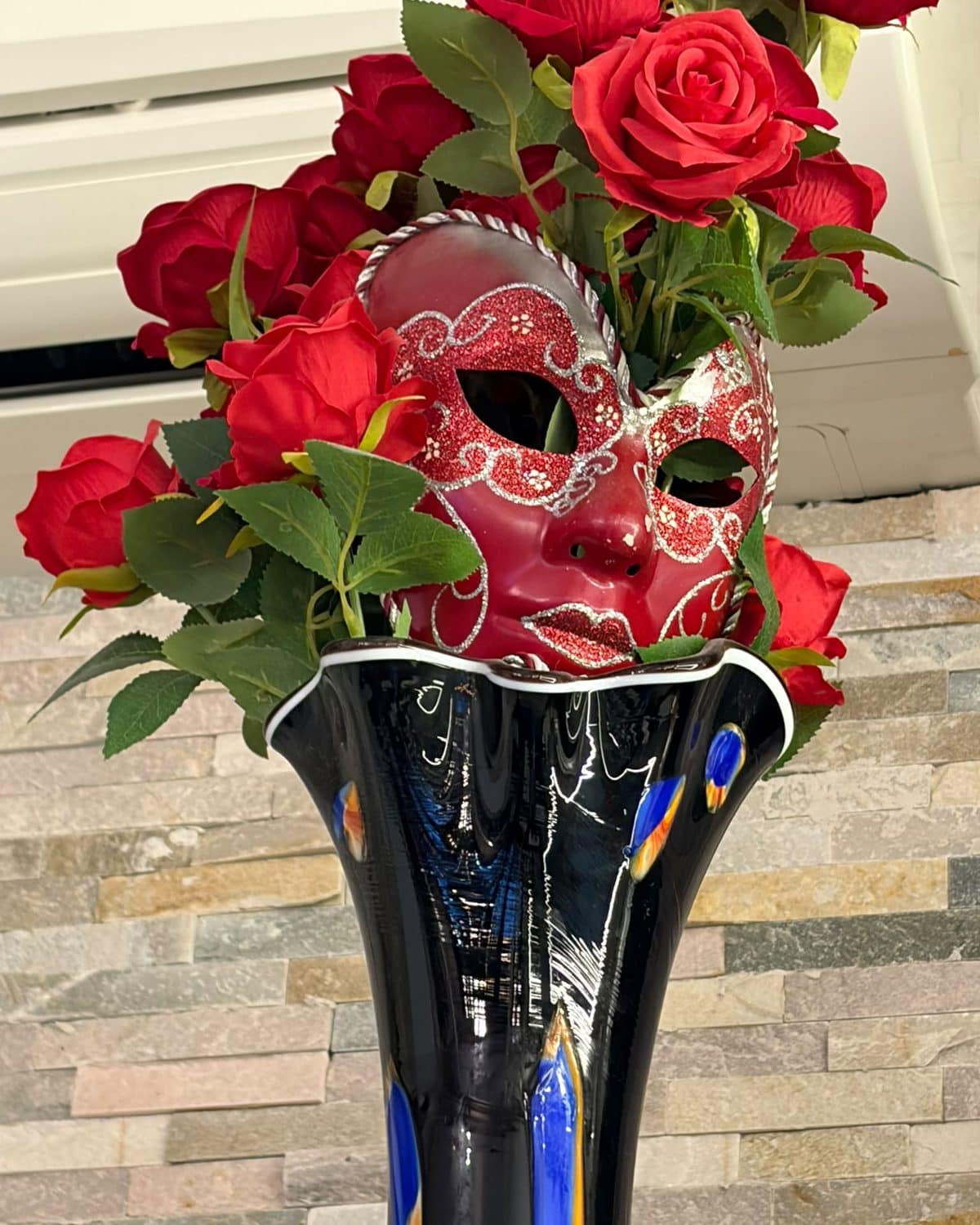 Masque vénitien rouge posé sur un vase de verre soufflé, entouré de roses.